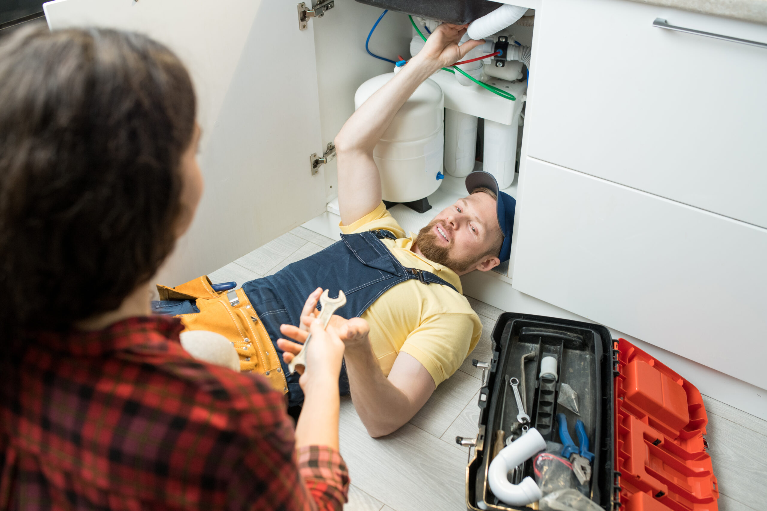 Smiling handsome young plumber in cap lying on floor in kitchen and fixing sink pipe while home owner giving wrench to him