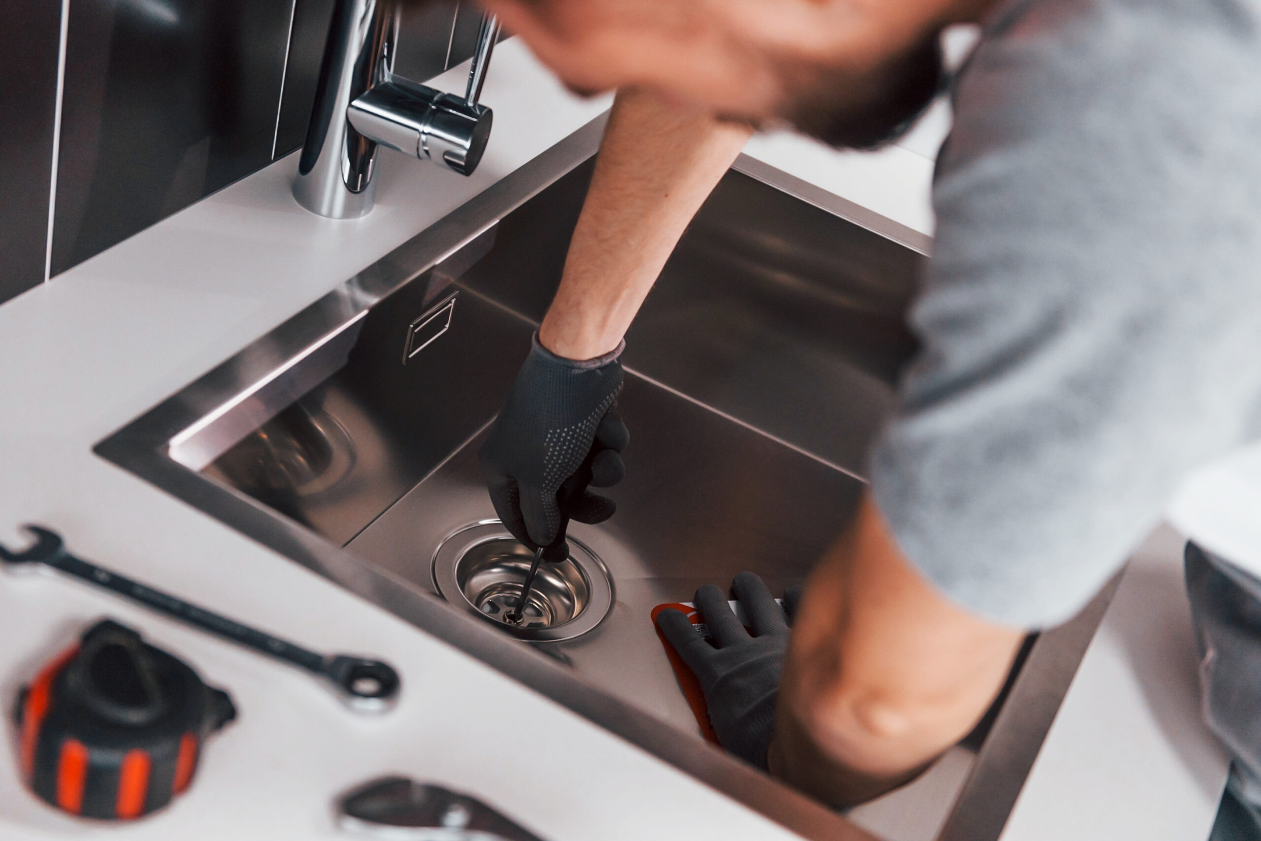 Young professional plumber in grey uniform working on the kitchen.